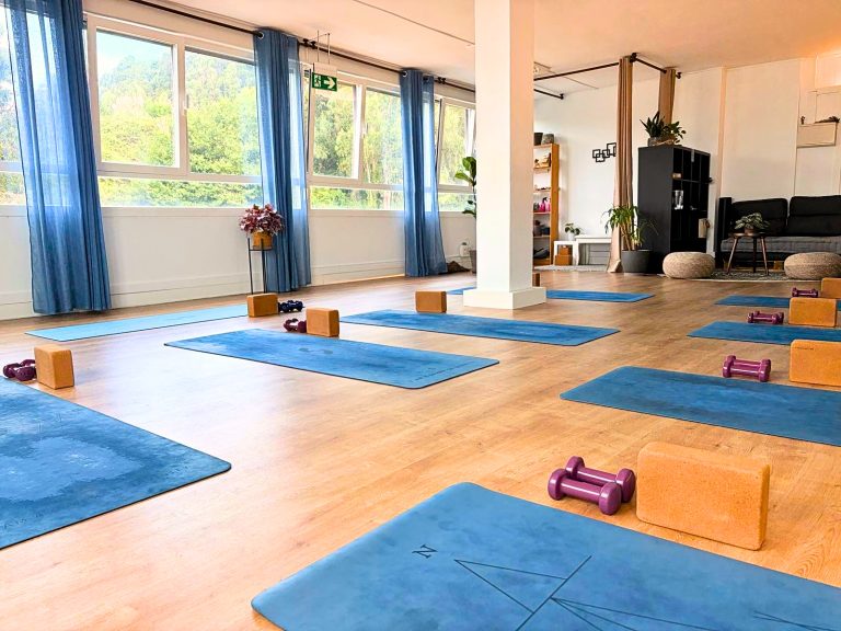 Sala de yoga luminosa con esterillas azules, bloques de corcho y mancuernas junto a grandes ventanales en Berango